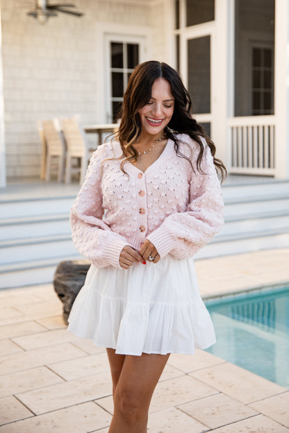 Woman wearing a pink cardigan and white skirt standing by a pool.