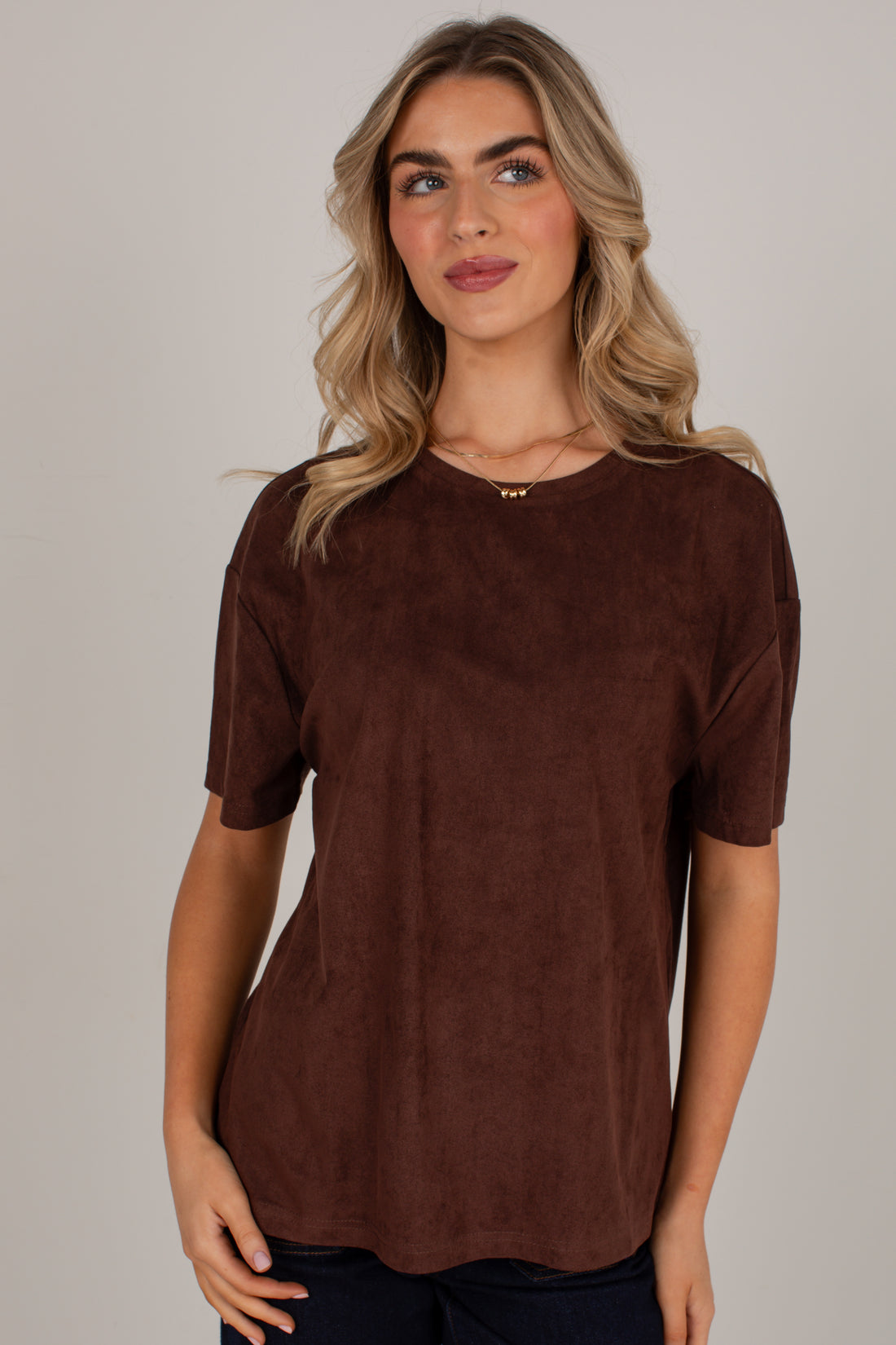 A woman wearing a short-sleeve crewneck top in a dark brown suede-like fabric, photographed against a plain light gray studio background.