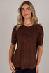 A woman wearing a short-sleeve crewneck top in a dark brown suede-like fabric, photographed against a plain light gray studio background.