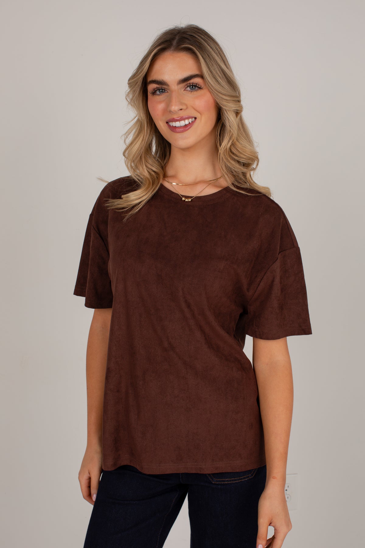A woman wearing a brown short-sleeve crewneck top with a textured suede-like finish, paired with dark pants, against a solid light gray background.