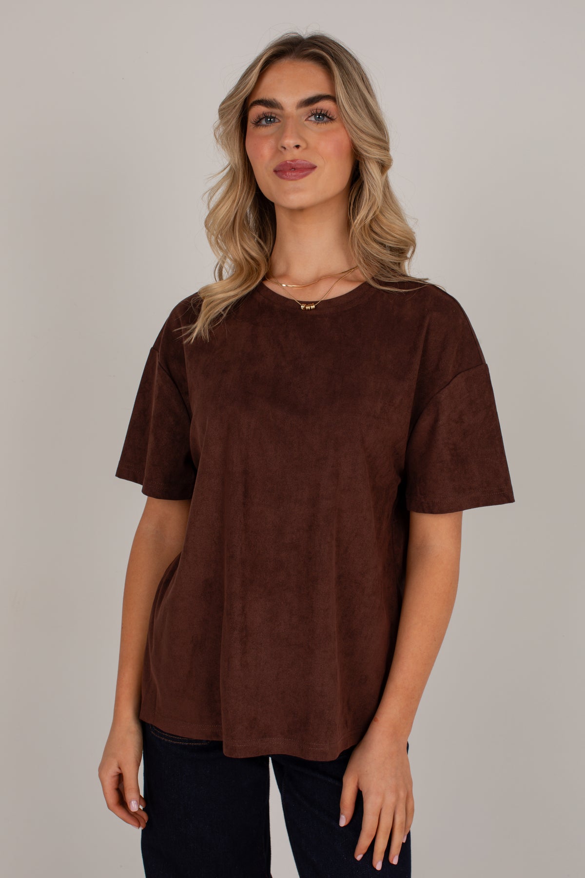 A woman modeling a brown short-sleeve top with a soft suede-like texture, paired with dark blue jeans, against a plain light gray studio background.