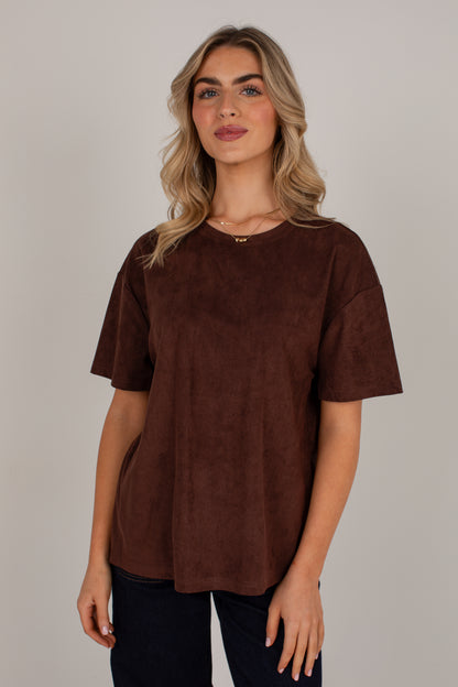A woman modeling a brown short-sleeve top with a soft suede-like texture, paired with dark blue jeans, against a plain light gray studio background.