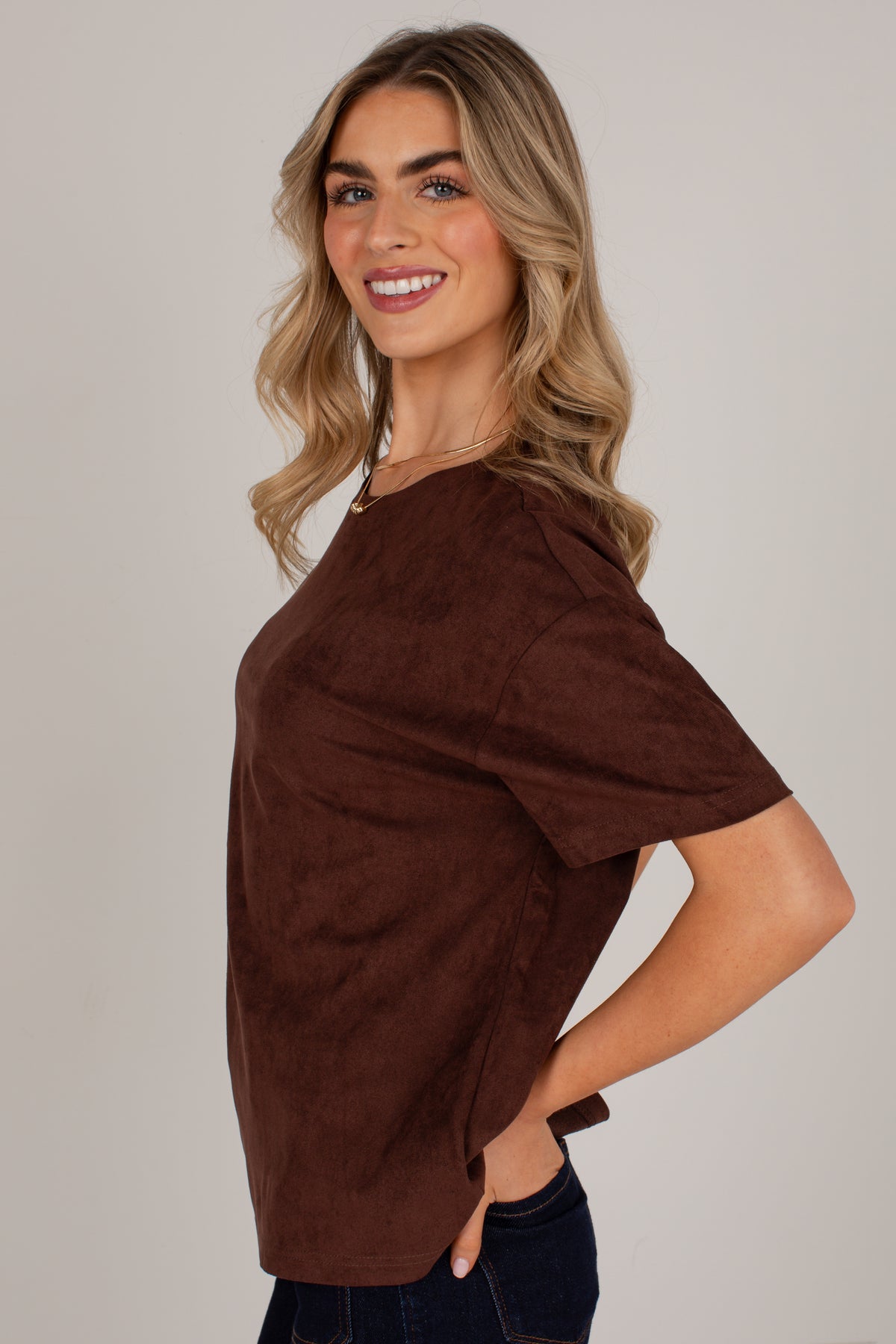 A woman modeling a brown short-sleeve top with a soft suede-like texture, paired with dark blue jeans, against a plain light gray studio background.