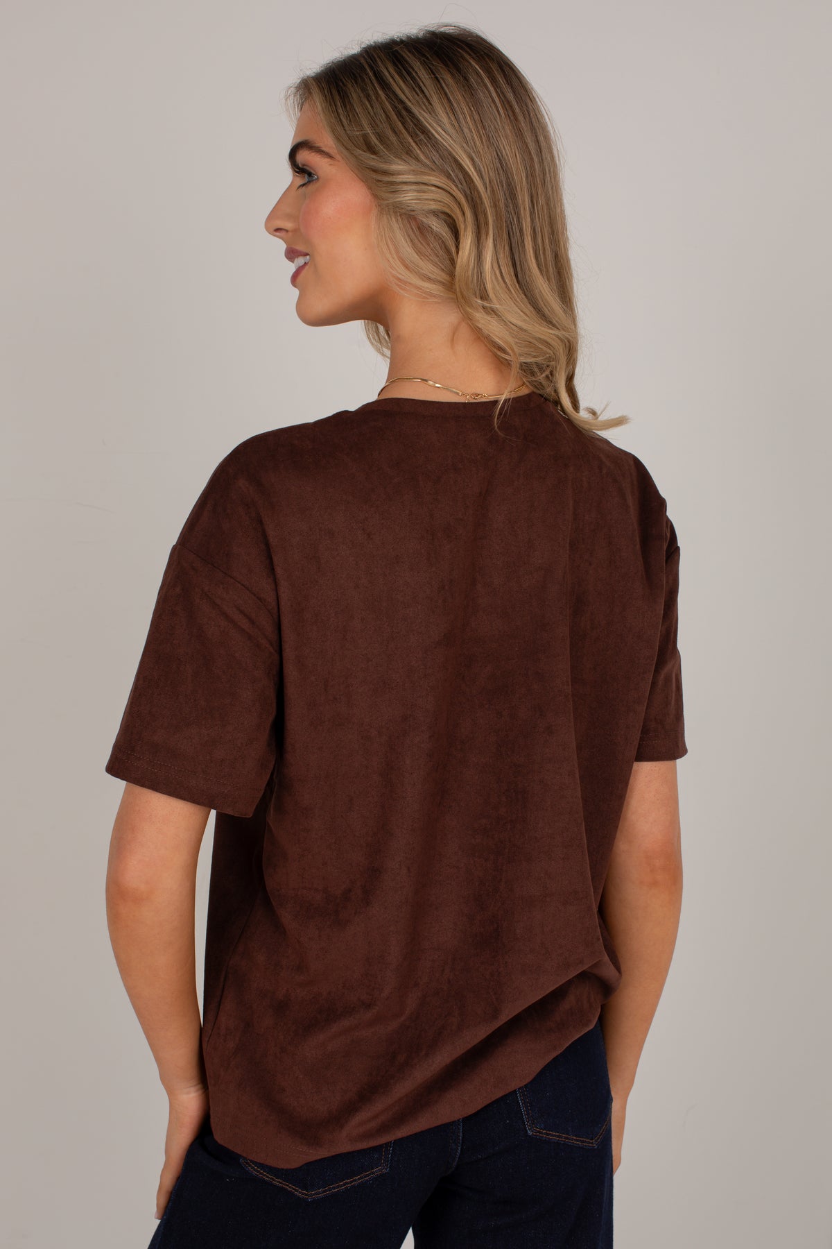 A woman shown from the back wearing an oversized short-sleeve brown top and dark blue jeans, photographed against a plain light gray studio background.