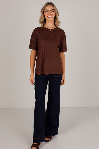 A woman modeling a brown short-sleeve V-neck top with dark navy wide-leg pants, standing against a plain light gray studio background.