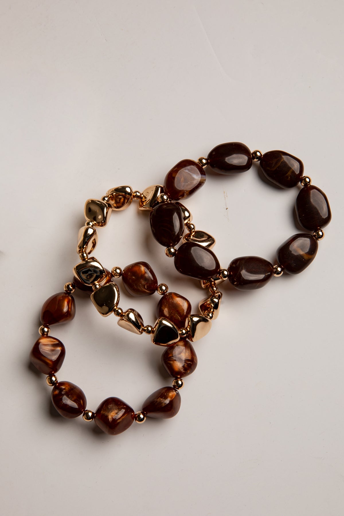 Two beaded bracelets with polished dark brown and amber stone-like beads separated by small gold-toned spacer beads, arranged on a light neutral background.
