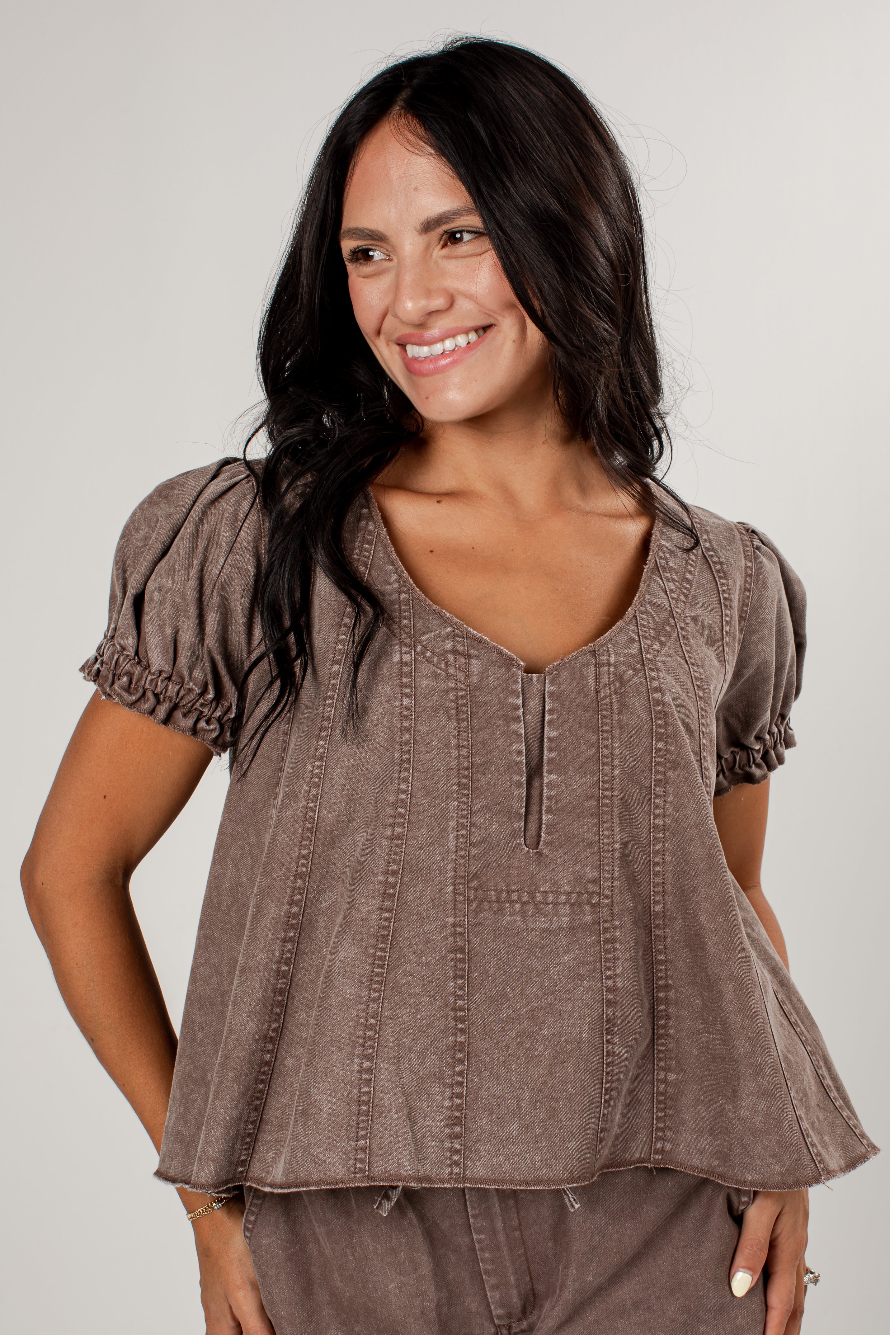A woman wearing a brown short-sleeve top with puff sleeves, a split V-neck, and vertical button details, paired with matching brown pants, against a light gray background.