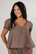A woman wearing a brown short-sleeve top with puff sleeves, a split V-neck, and vertical button details, paired with matching brown pants, against a light gray background.