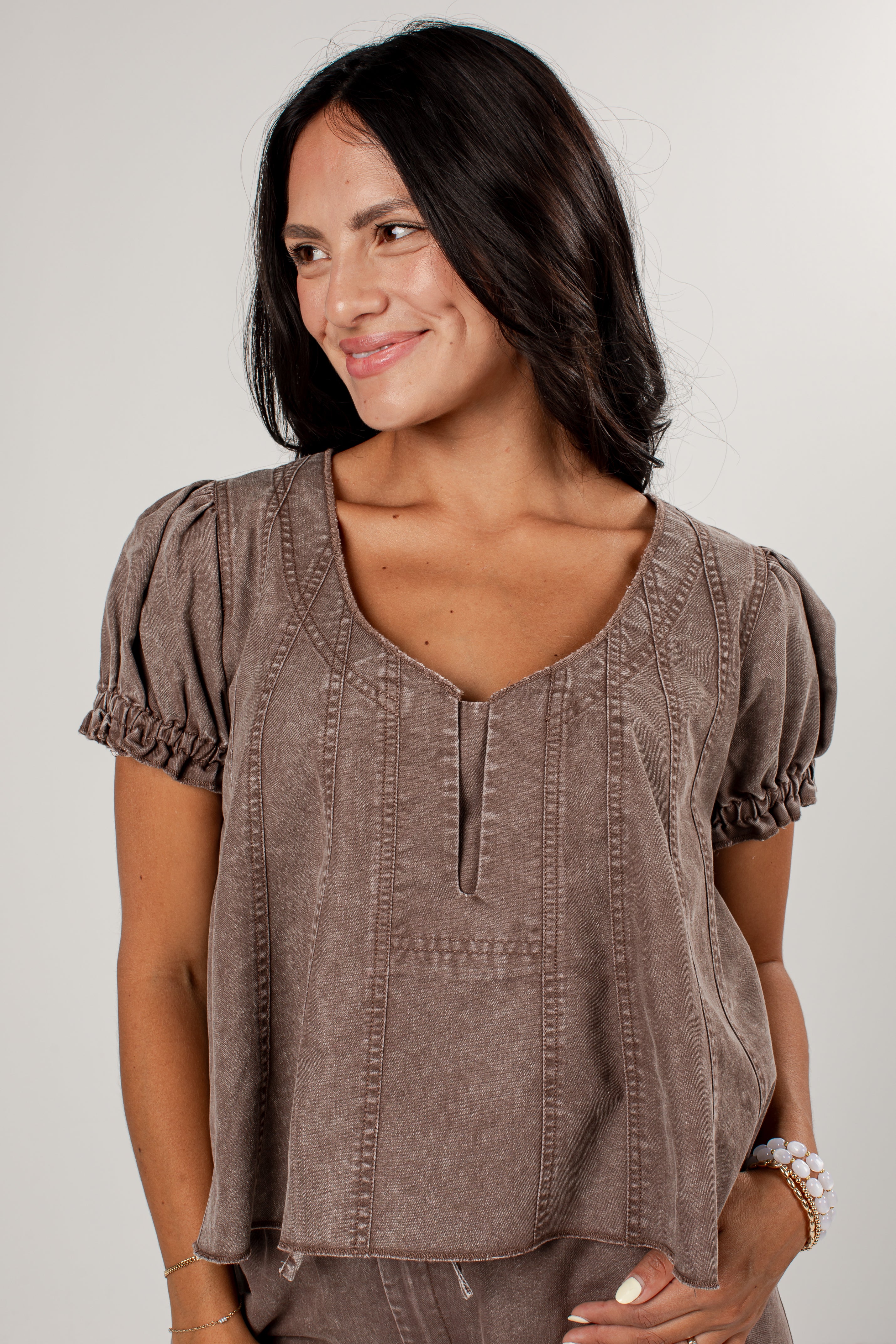 A woman wearing a taupe brown short-sleeve top with puff sleeves, a split neckline, and vertical stitched details, photographed against a light gray background.