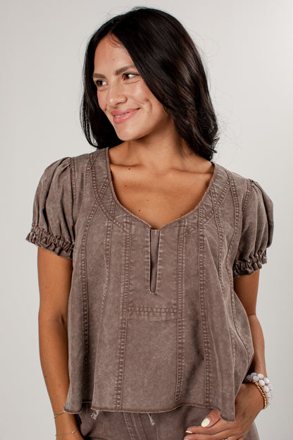A woman wearing a taupe brown short-sleeve top with puff sleeves, a split neckline, and vertical stitched details, photographed against a light gray background.