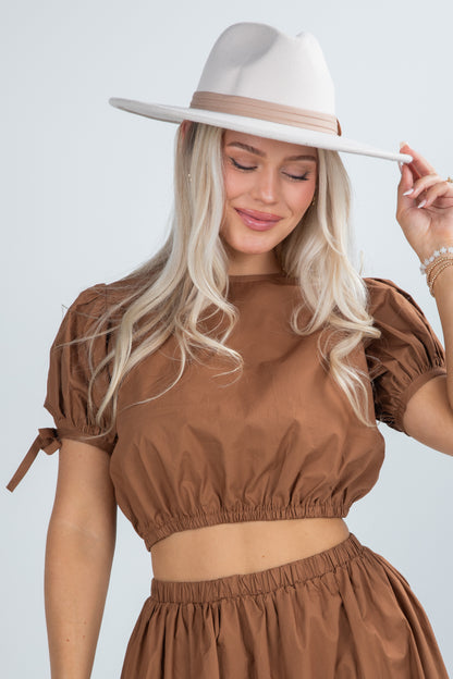Woman wearing a brown outfit and white hat against a light gray background