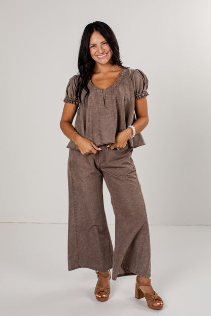 A woman modeling a matching two-piece outfit consisting of a short-sleeve cropped top with puff sleeves and wide-leg pants, both in a brown linen-like fabric, paired with brown heeled sandals against a plain light gray studio background.