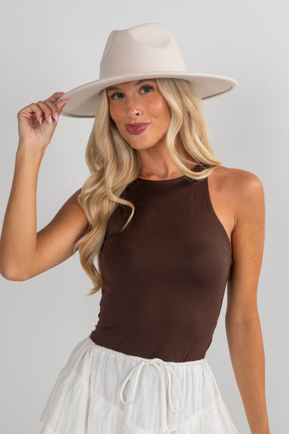 Woman wearing a beige hat and brown top with a white skirt on a gray background