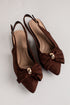 A pair of dark brown suede slingback pointed-toe flats with a large bow on the toe and a small gold-tone metal accent, displayed on a light gray background.