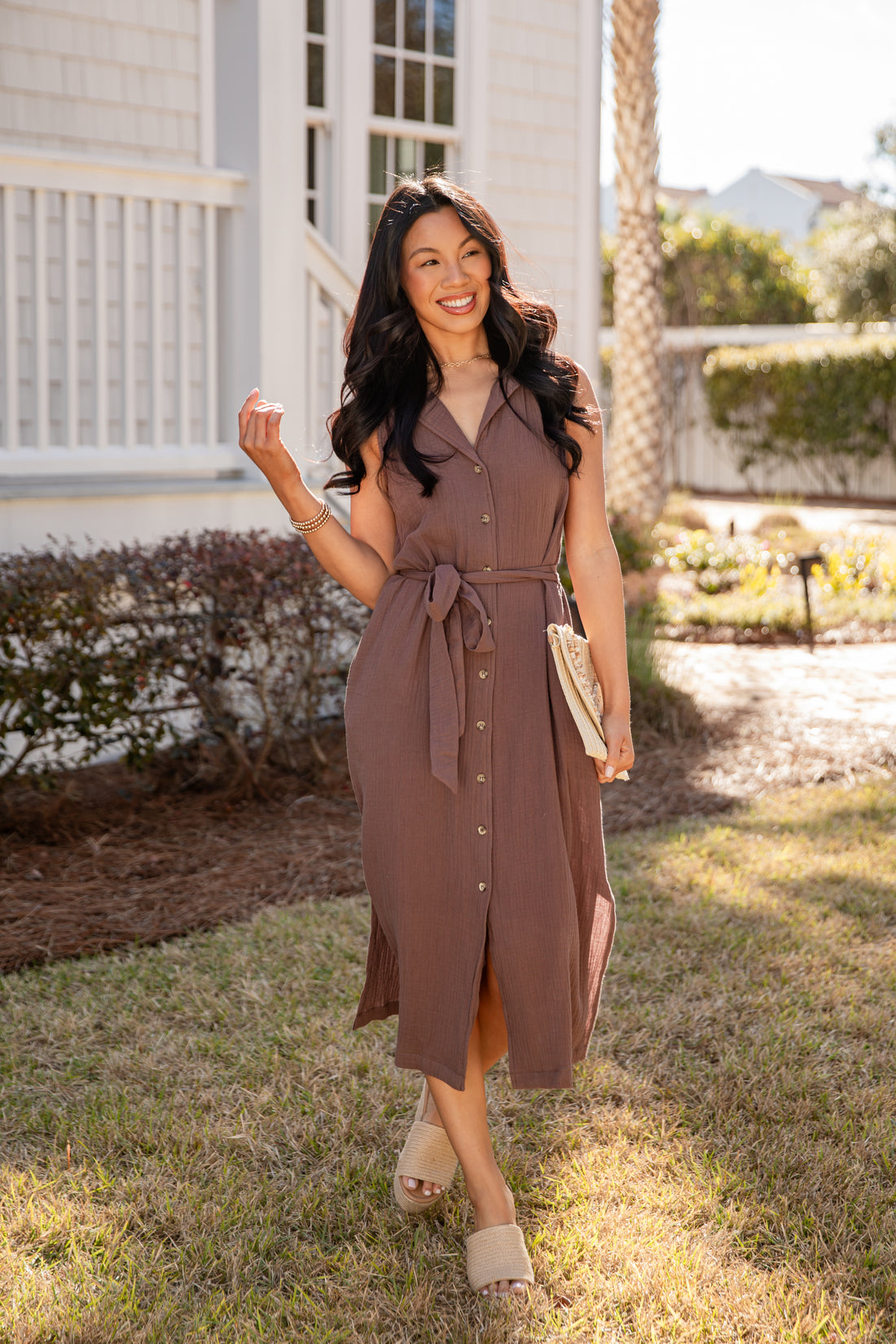 Woman in a brown dress standing outdoors near a house with a palm tree.