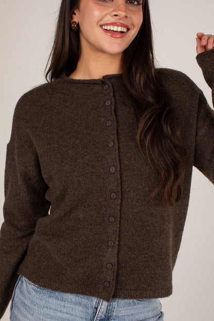 A woman wearing a dark brown button-up knit cardigan sweater and light blue jeans, smiling against a plain light beige background.
