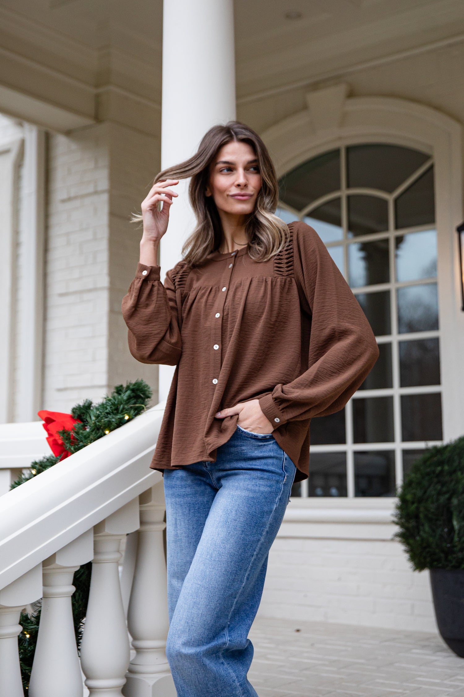 Woman in a brown blouse and blue jeans standing on a porch with decorative elements.