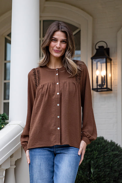 Woman wearing a brown blouse and blue jeans standing on a porch.