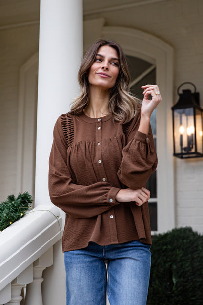 Woman wearing a brown blouse and blue jeans standing on a porch.