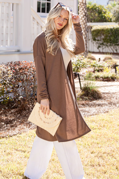 Woman in a brown cardigan and white pants standing outdoors.