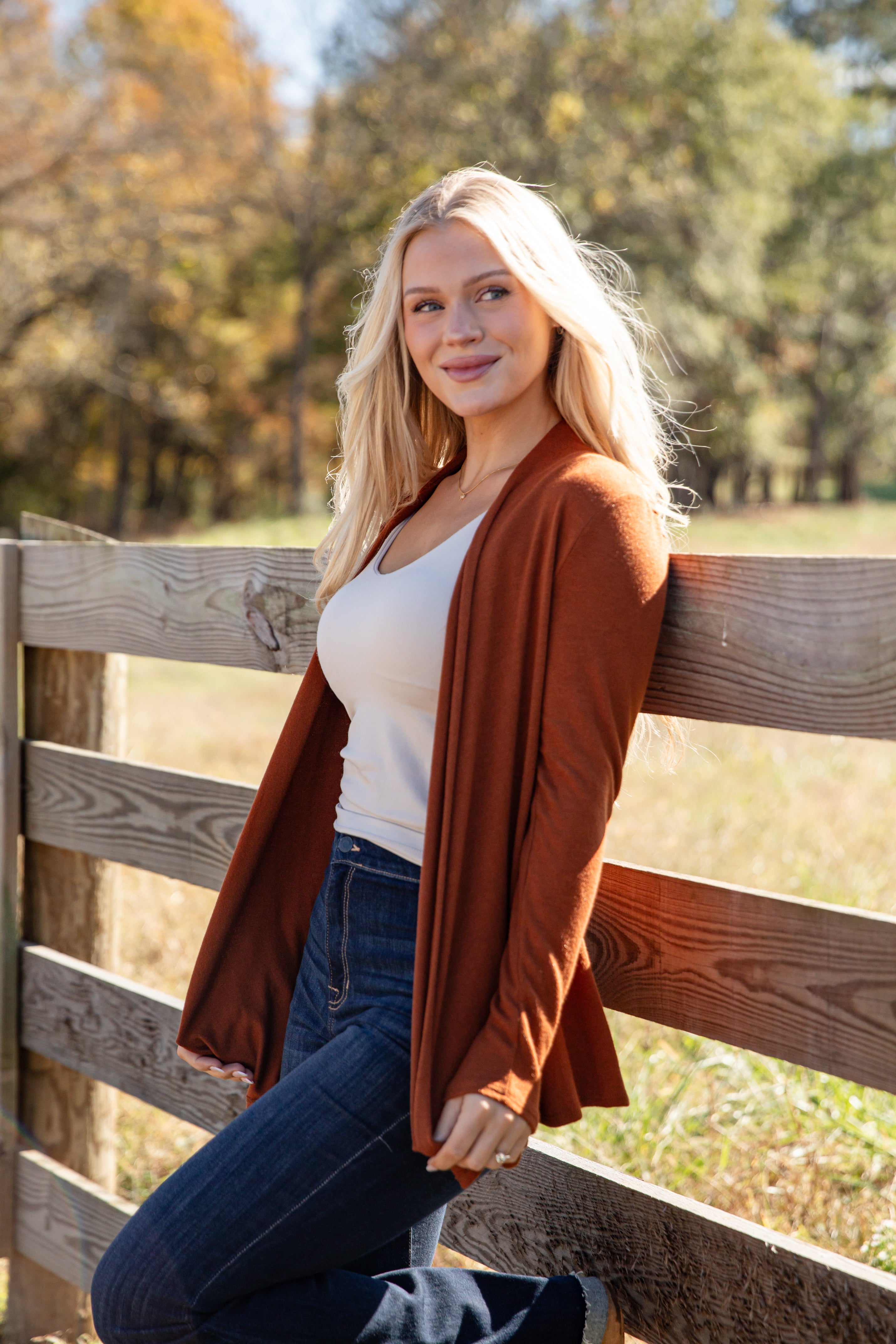 Woman wearing a brown cardigan over a white top and blue jeans, standing in front of a wooden fence with trees in the background.