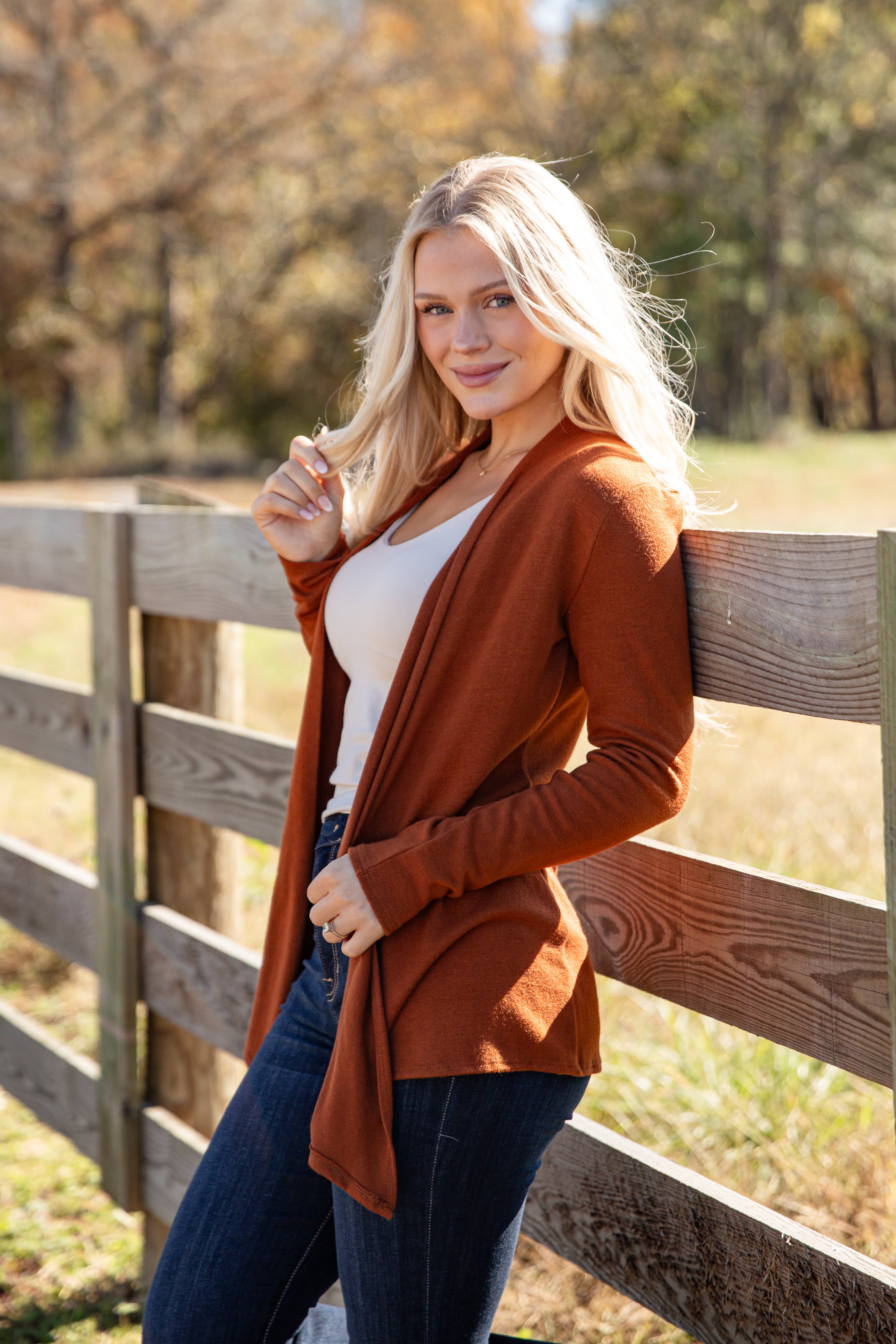 Woman wearing a brown cardigan over a white top and blue jeans, standing in front of a wooden fence with trees in the background.