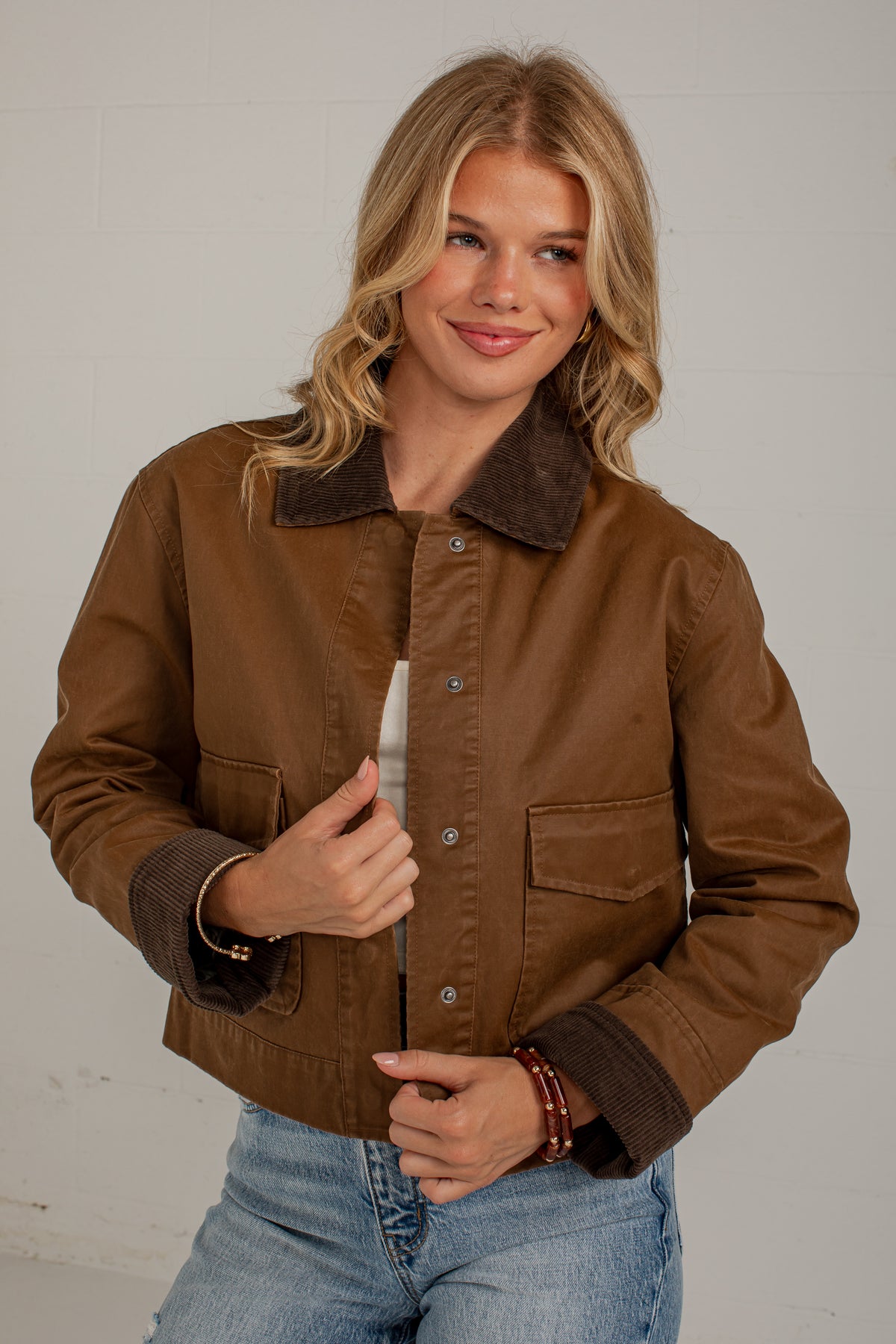 A woman wearing a brown jacket with a dark brown corduroy collar and cuffs, paired with light blue jeans, standing against a light gray wall.