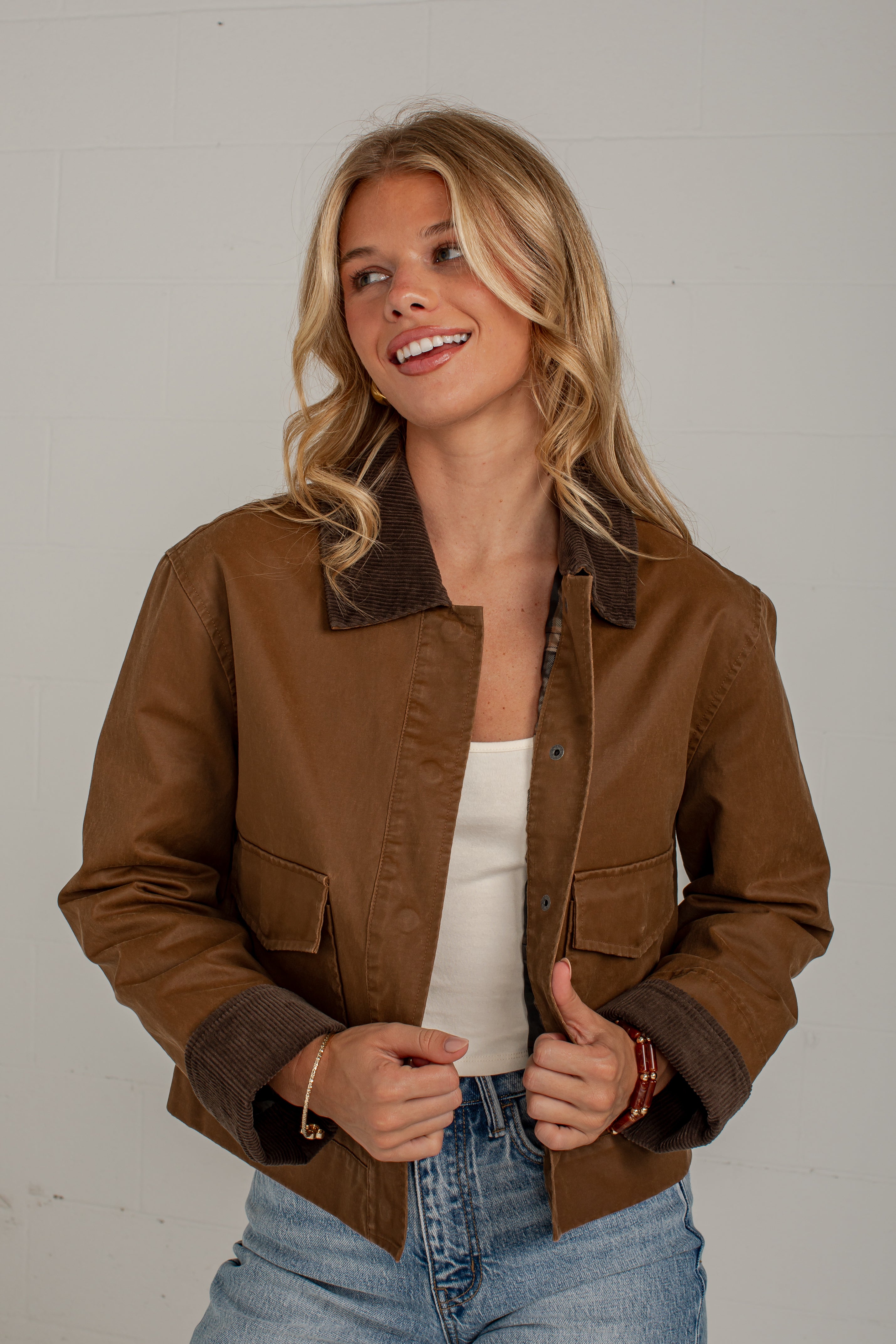 A woman wearing a brown faux-leather bomber jacket with a dark brown fleece collar and cuffs, styled with a white tank top and blue jeans against a light gray wall.