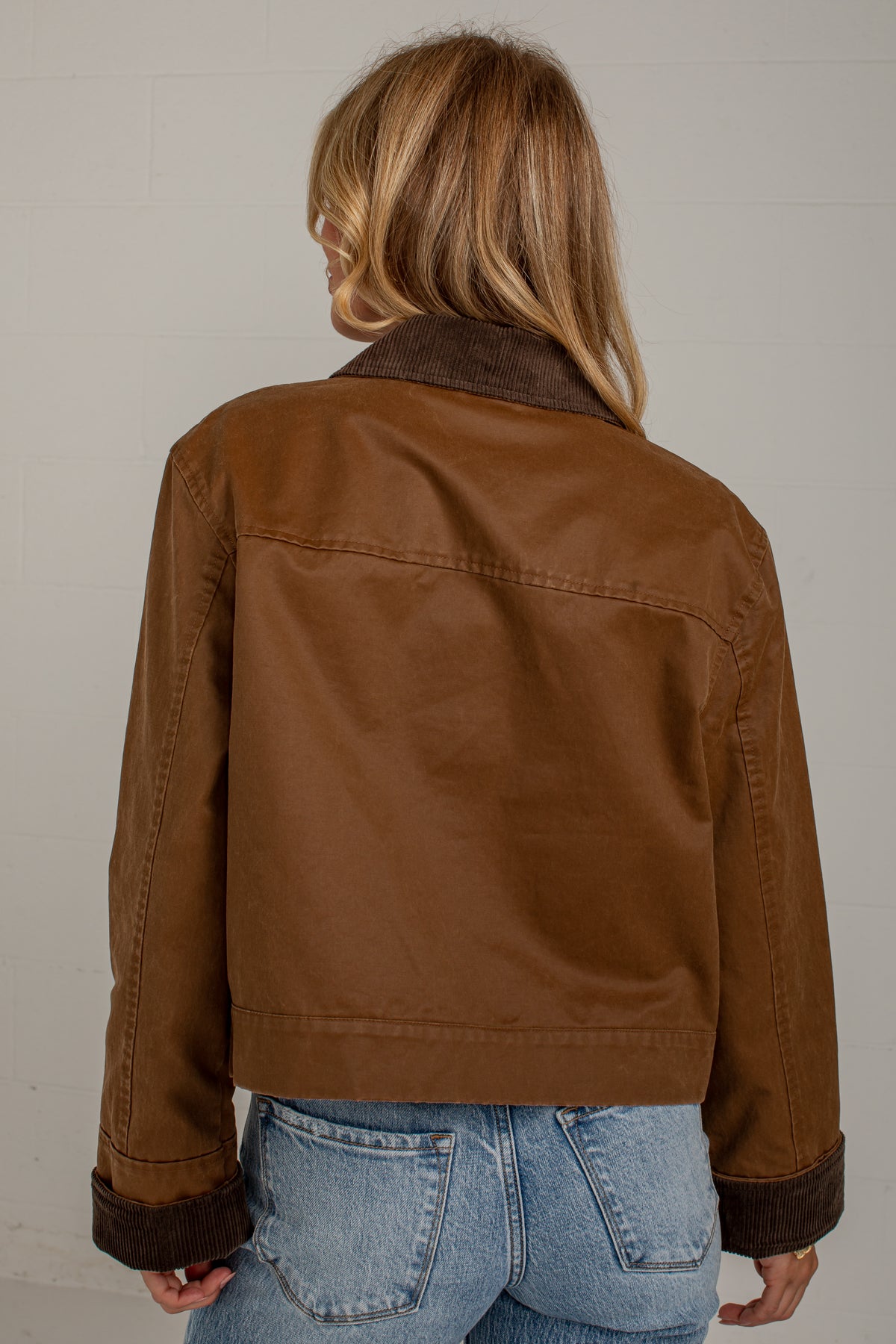 A woman viewed from behind wearing a cropped brown jacket with dark brown corduroy collar and cuffs, paired with light blue jeans, against a light gray brick wall.