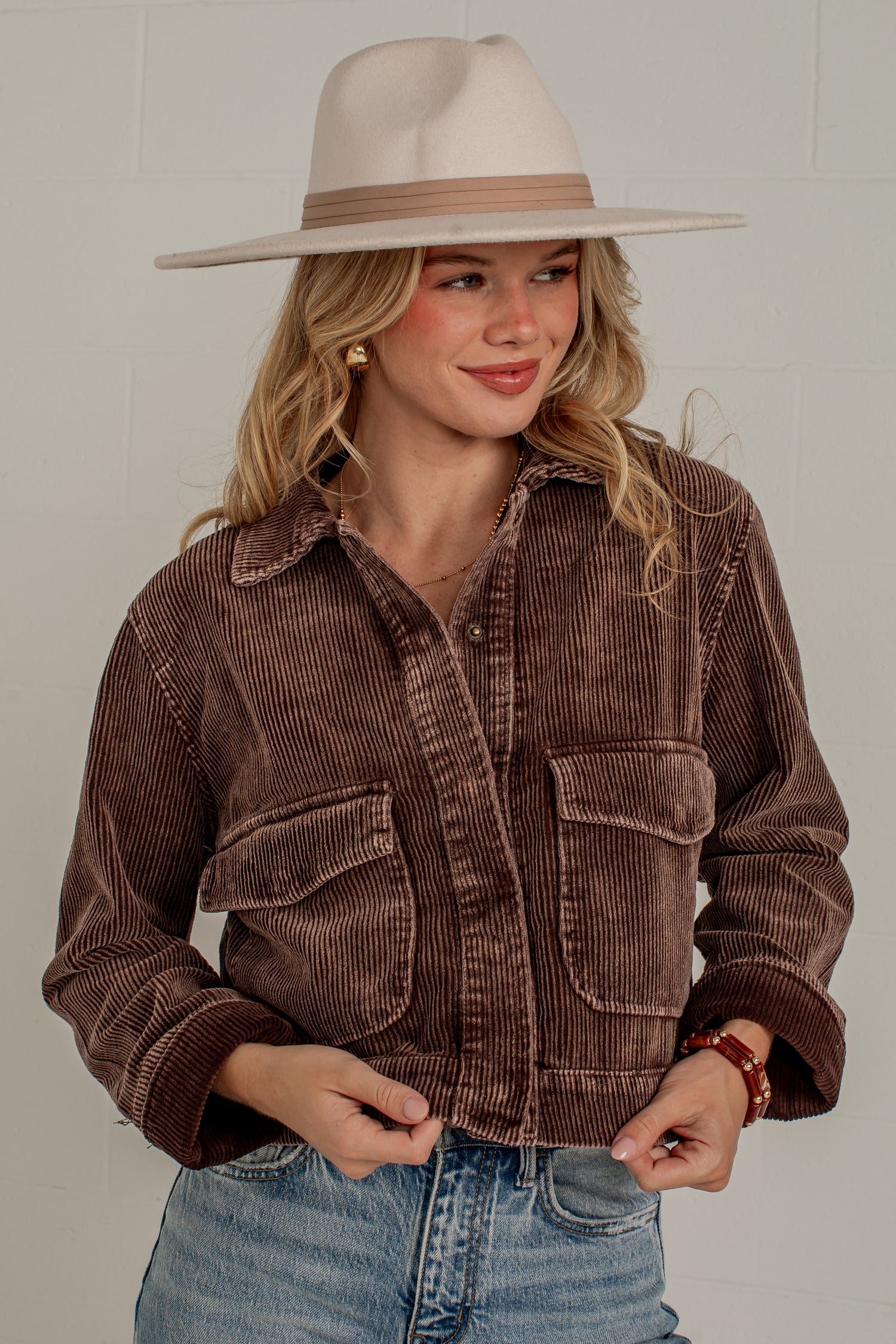 A woman wearing a brown corduroy button-up jacket, blue jeans, and a beige wide-brim hat, standing against a light gray brick wall.