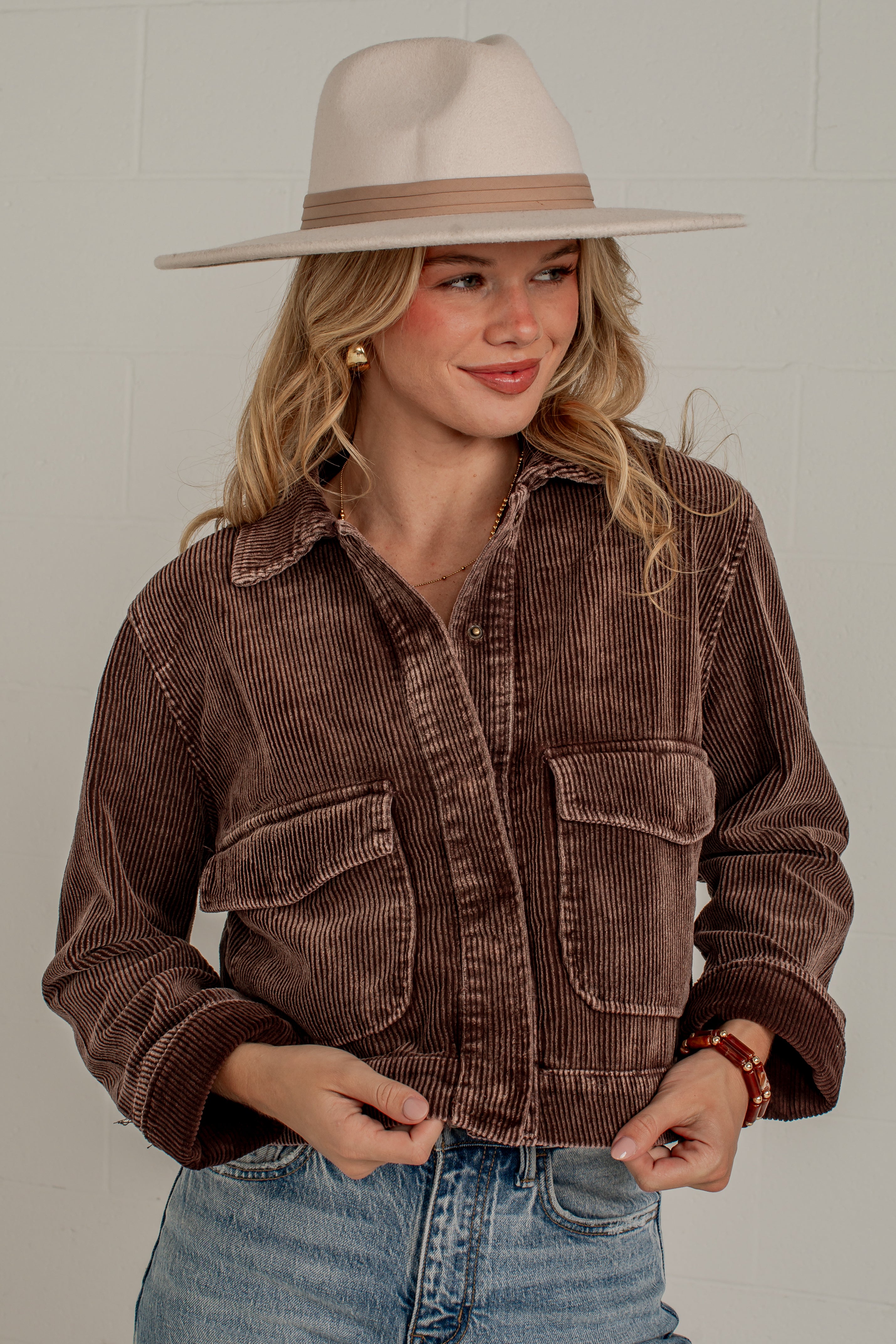 A woman wearing a brown corduroy button-up jacket, blue jeans, and a beige wide-brim hat, standing against a light gray brick wall.