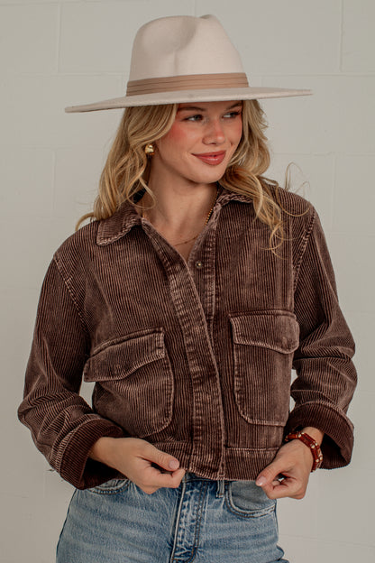 A woman wearing a brown corduroy button-up jacket, blue jeans, and a beige wide-brim hat, standing against a light gray brick wall.