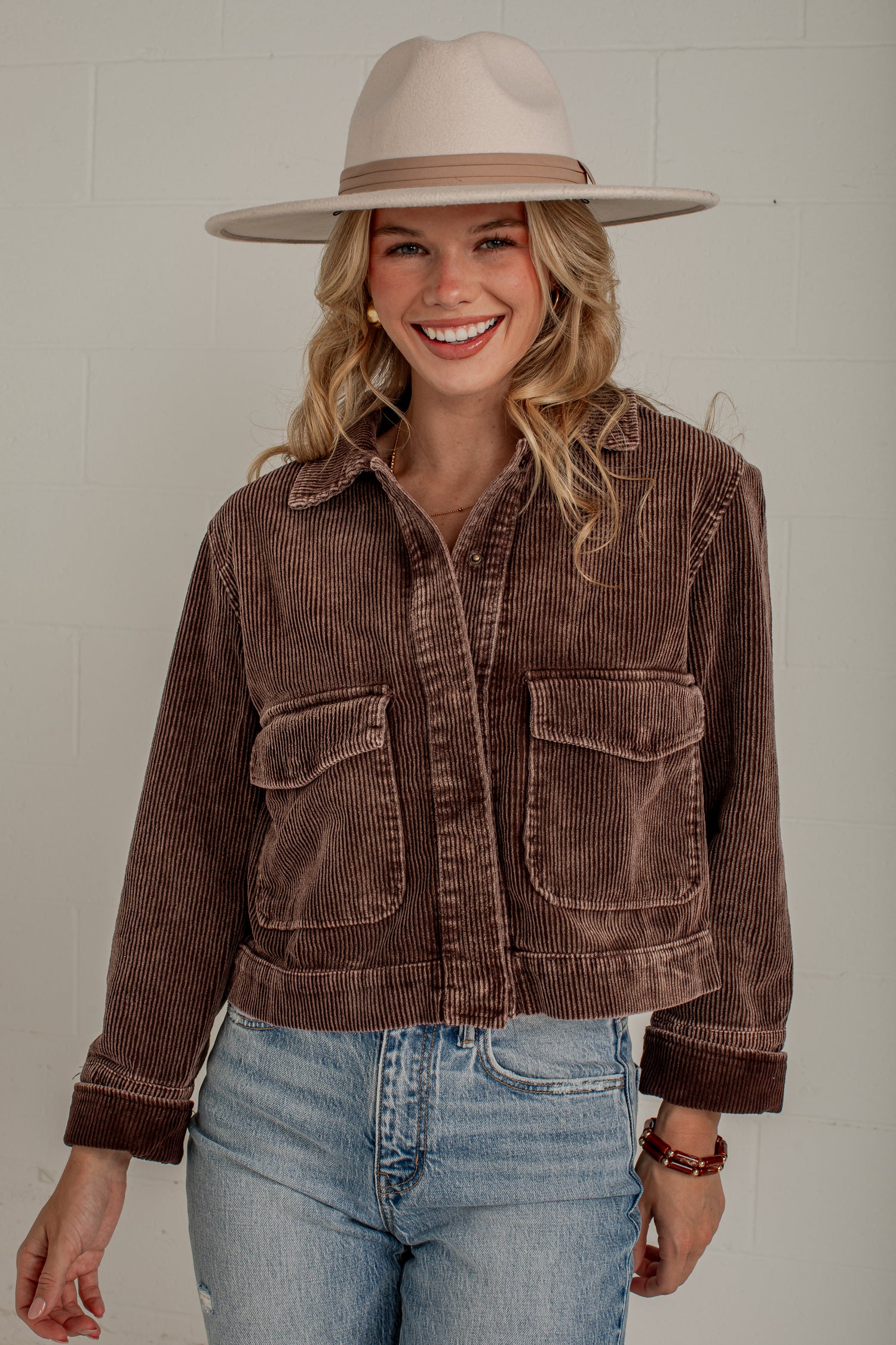 A woman wearing a brown corduroy cropped jacket with front flap pockets, paired with light blue jeans and a beige wide-brim hat, standing against a light gray wall.