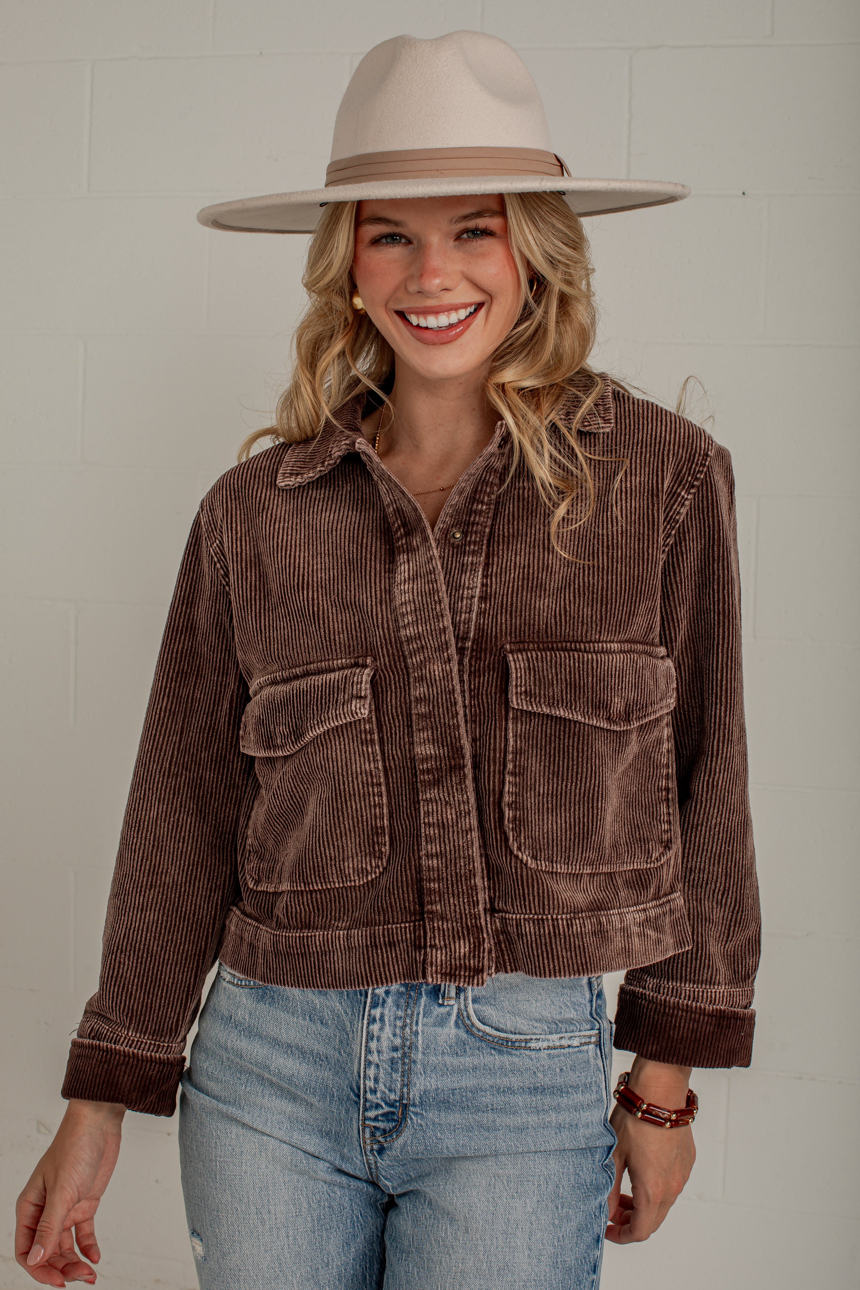 A woman wearing a brown corduroy cropped jacket with front flap pockets, paired with light blue jeans and a beige wide-brim hat, standing against a light gray wall.