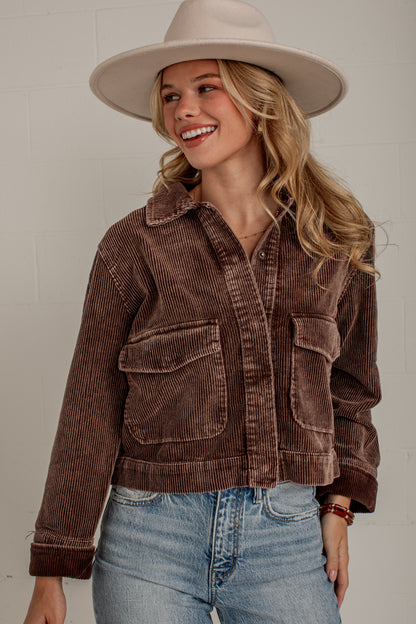 A woman wearing a brown corduroy jacket, blue jeans, and a beige wide-brim hat, smiling against a light gray brick wall.