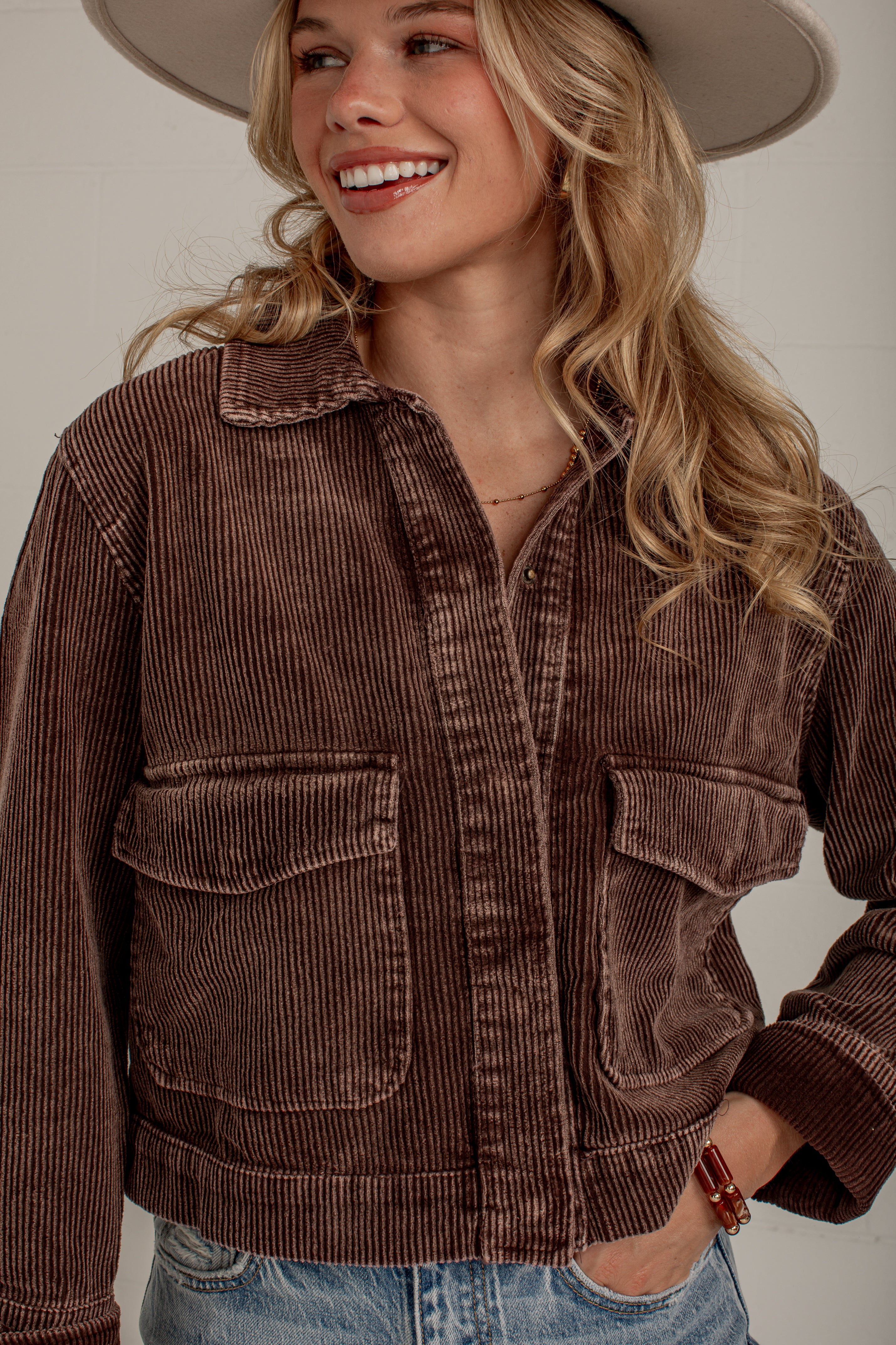A woman wearing a brown corduroy jacket with front flap pockets, styled with blue jeans and a wide-brim hat against a neutral beige background.