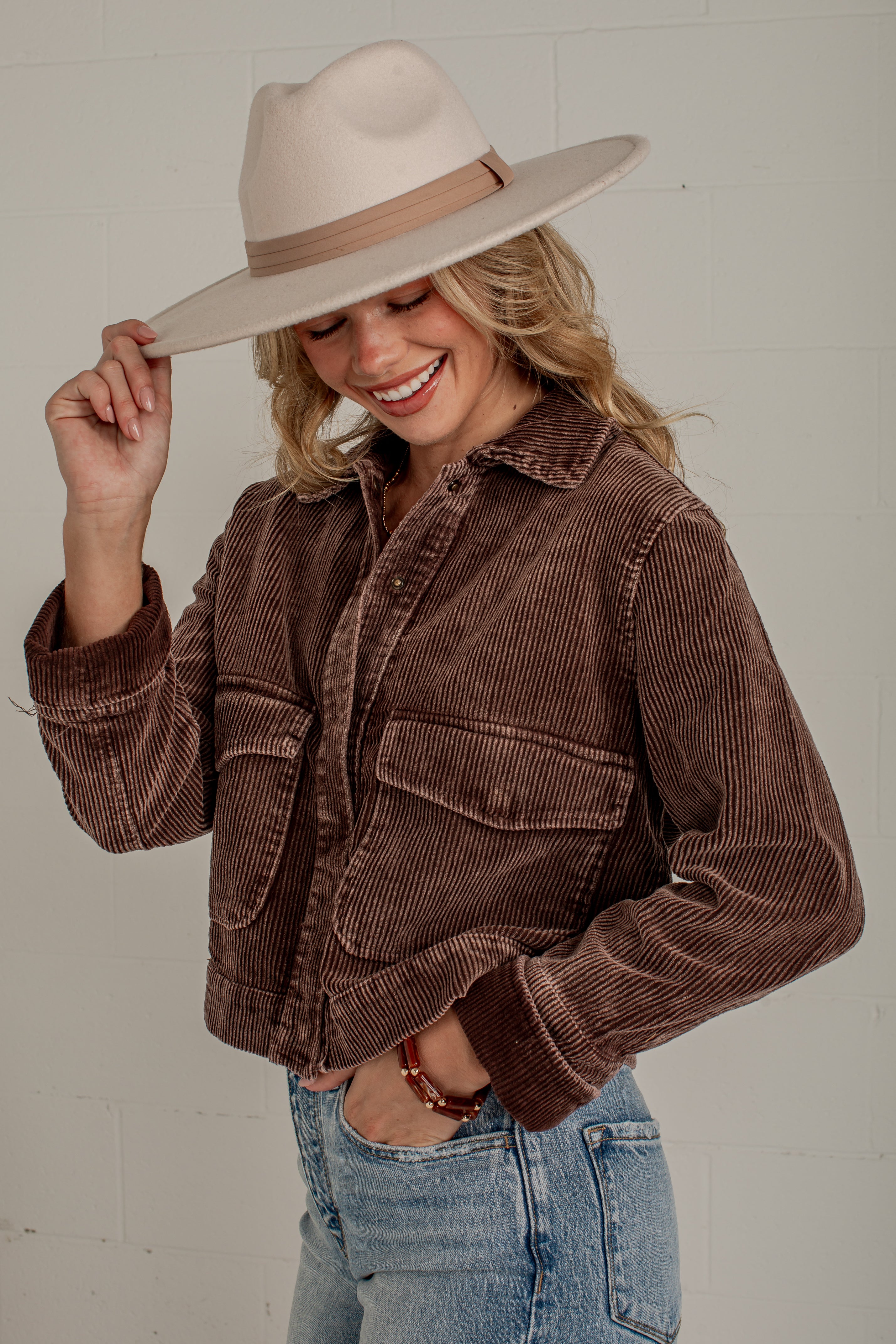 A woman wearing a brown corduroy jacket, light blue jeans, and a beige wide-brim hat, smiling and posing against a white brick wall.