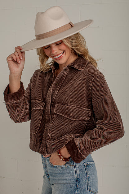 A woman wearing a brown corduroy jacket, light blue jeans, and a beige wide-brim hat, smiling and posing against a white brick wall.