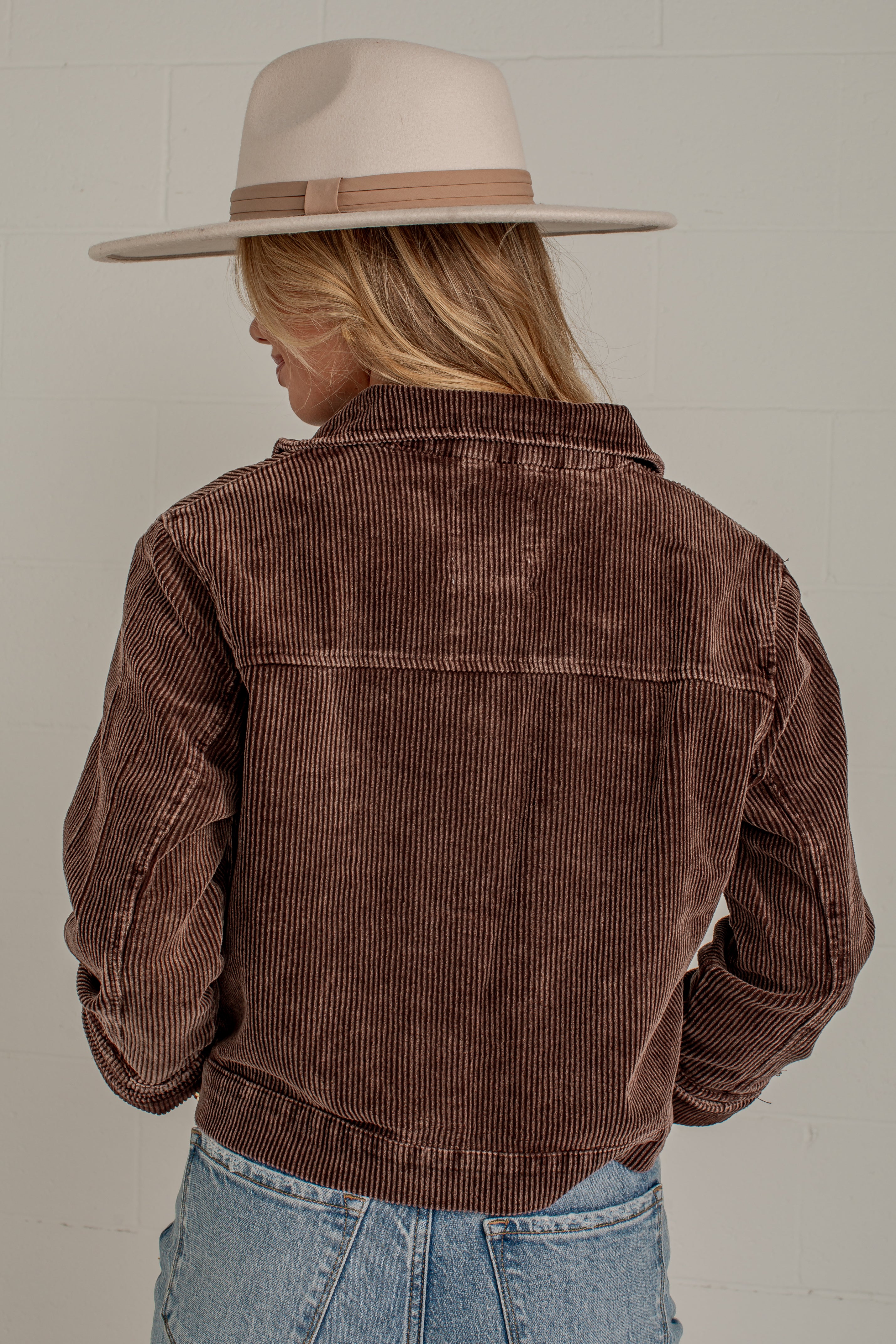 A woman shown from the back wearing a brown corduroy jacket, a beige wide-brim hat, and light blue jeans against a light gray brick wall.