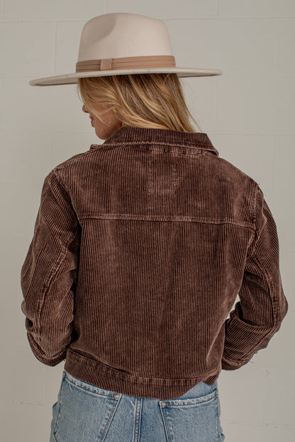 A woman shown from the back wearing a brown corduroy jacket, a beige wide-brim hat, and light blue jeans against a light gray brick wall.