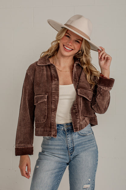 A woman wearing a brown corduroy cropped jacket, a cream tank top, light blue jeans, and a beige wide-brim hat, posing against a light gray brick wall.