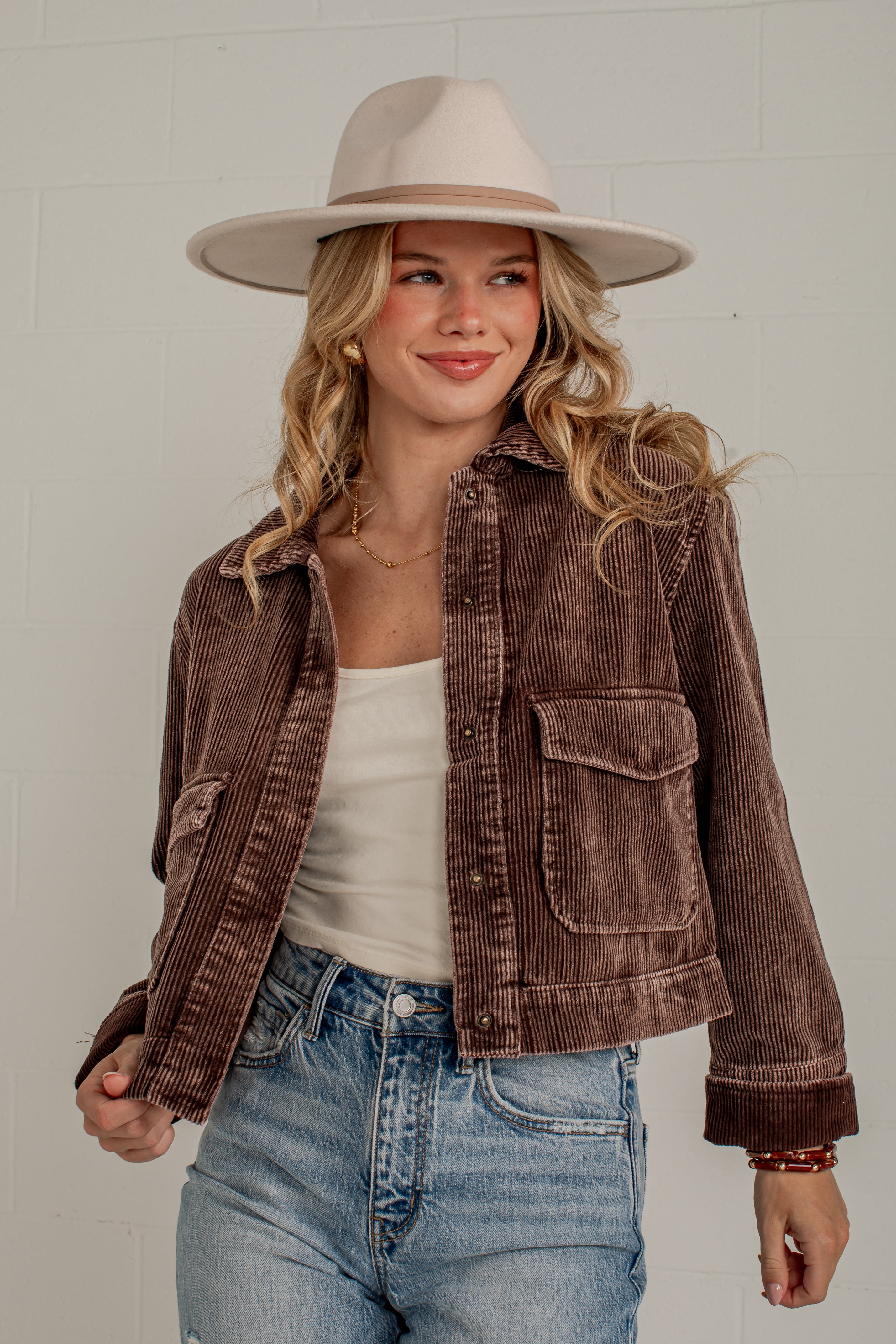 A woman wearing a brown corduroy jacket, a beige wide-brim hat, a white tank top, and blue jeans, standing against a light gray brick wall.