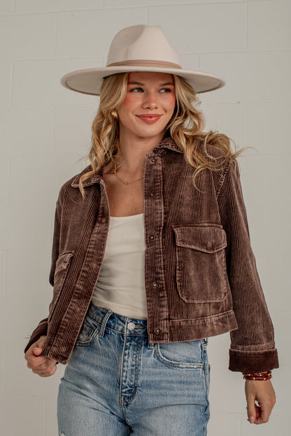A woman wearing a brown corduroy jacket, a beige wide-brim hat, a white tank top, and blue jeans, standing against a light gray brick wall.
