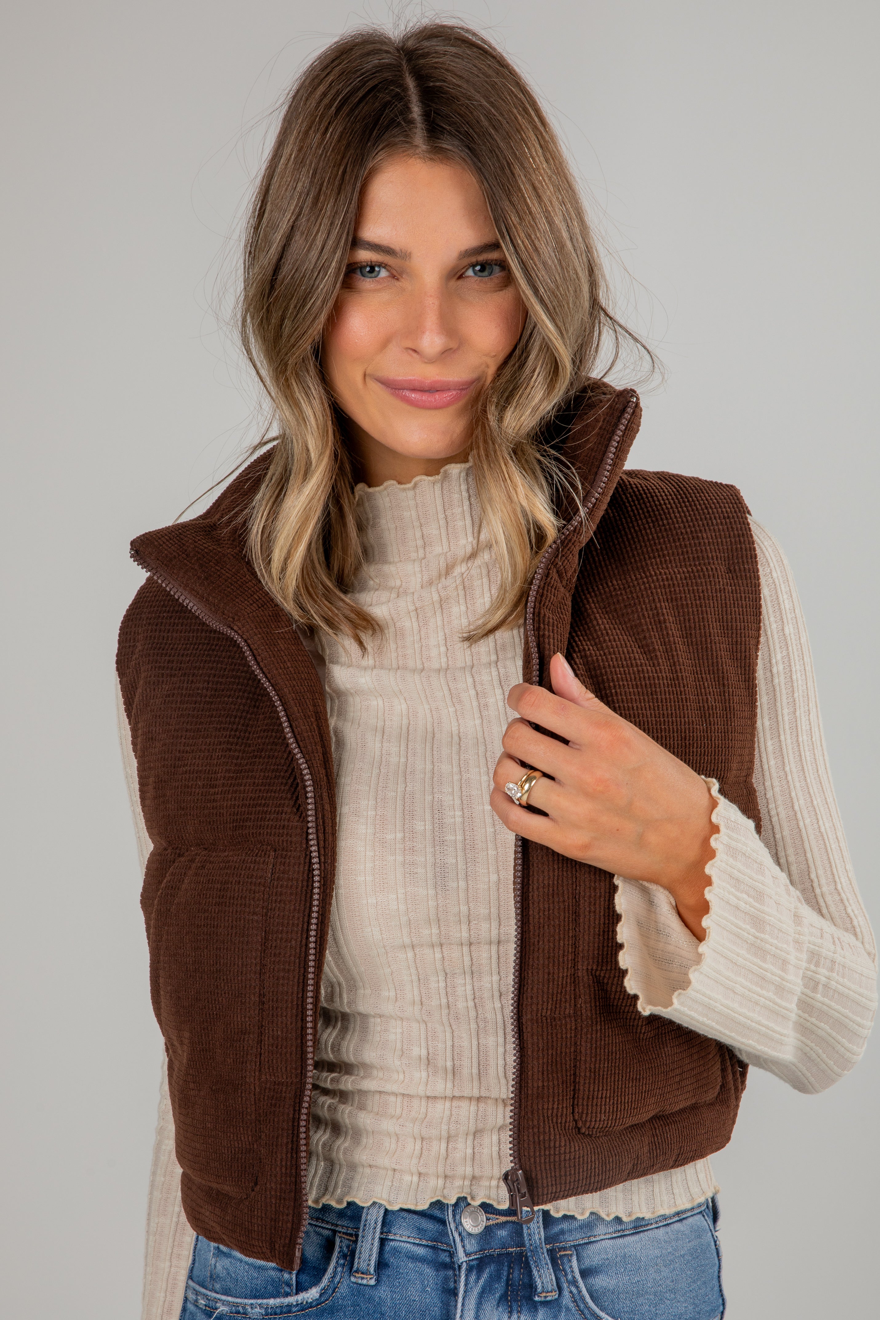 Woman wearing a brown vest over a beige sweater with a plain background