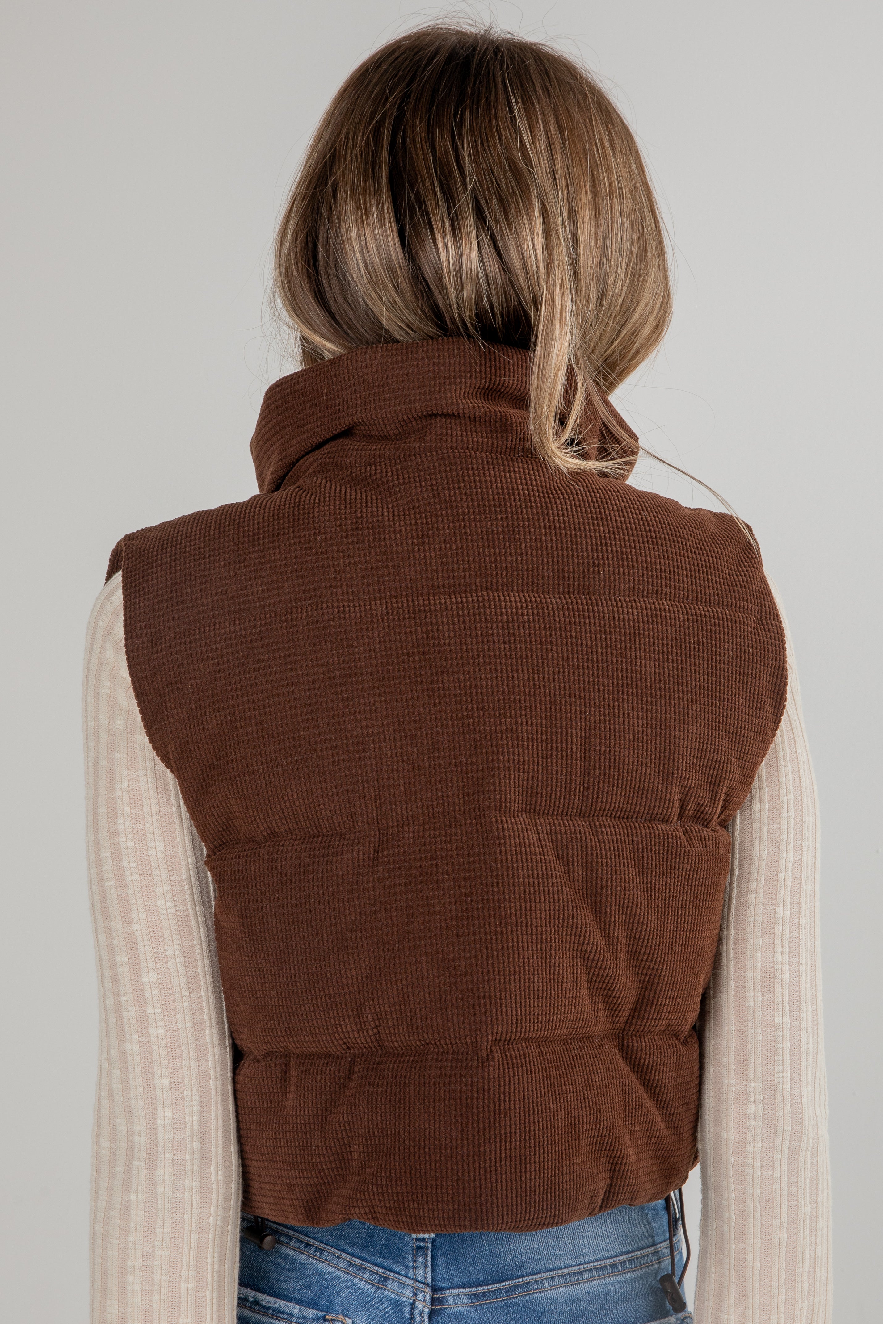 Person wearing a brown corduroy vest over a beige sweater and blue jeans on a plain background