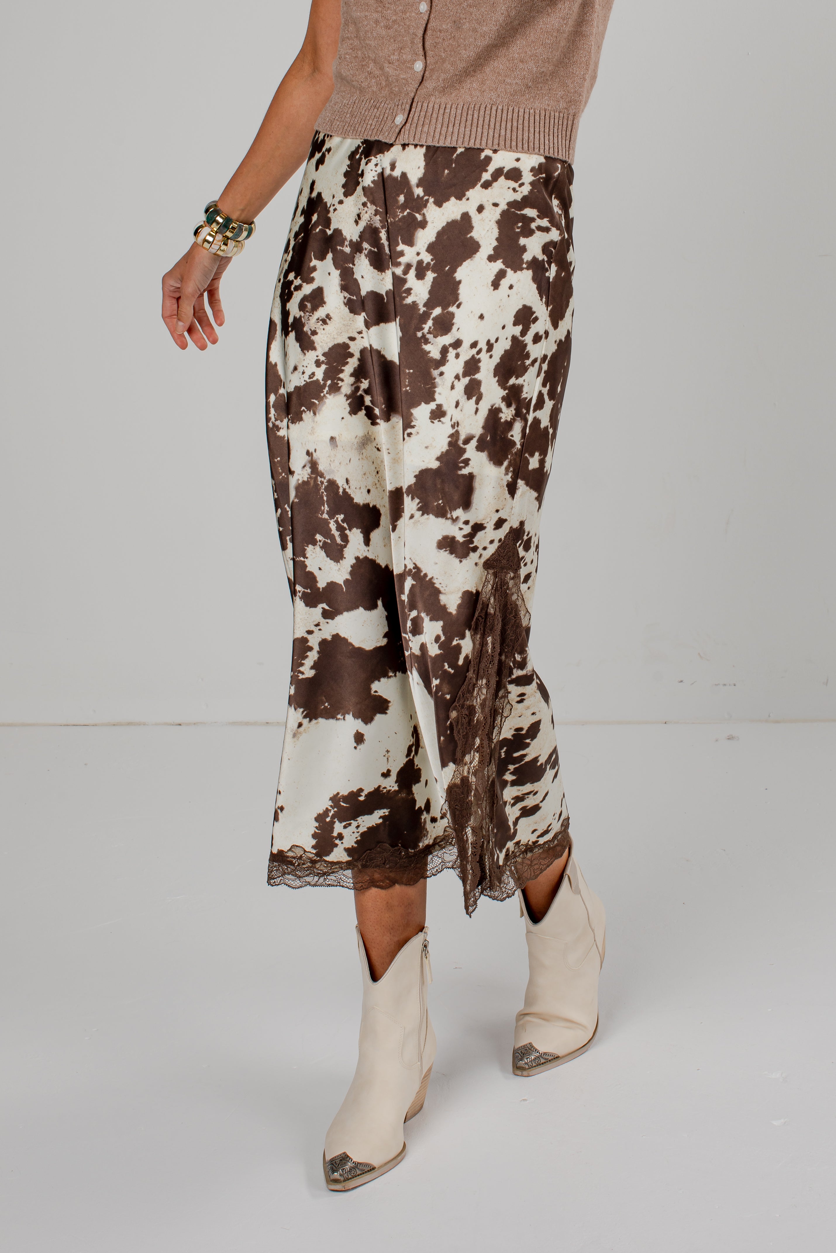 A model is shown from the waist down wearing a brown and cream cow-print skirt with a lace hem, paired with beige ankle boots and a taupe cardigan, against a plain light gray studio background.