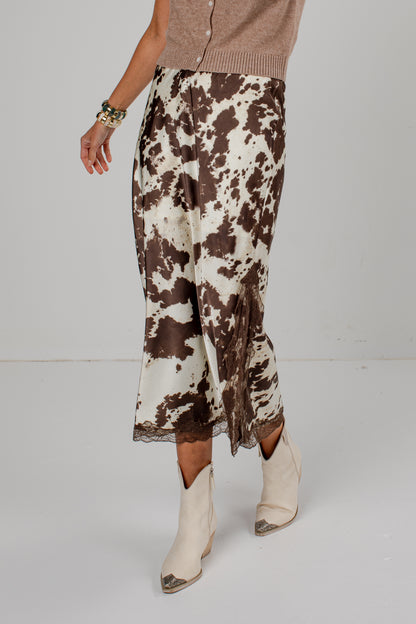 A model is shown from the waist down wearing a brown and cream cow-print skirt with a lace hem, paired with beige ankle boots and a taupe cardigan, against a plain light gray studio background.