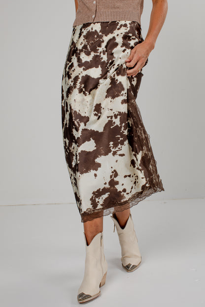 A model is shown from the waist down wearing a brown and white cow-print midi skirt with a lace hem, paired with cream-colored pointed-toe ankle boots.