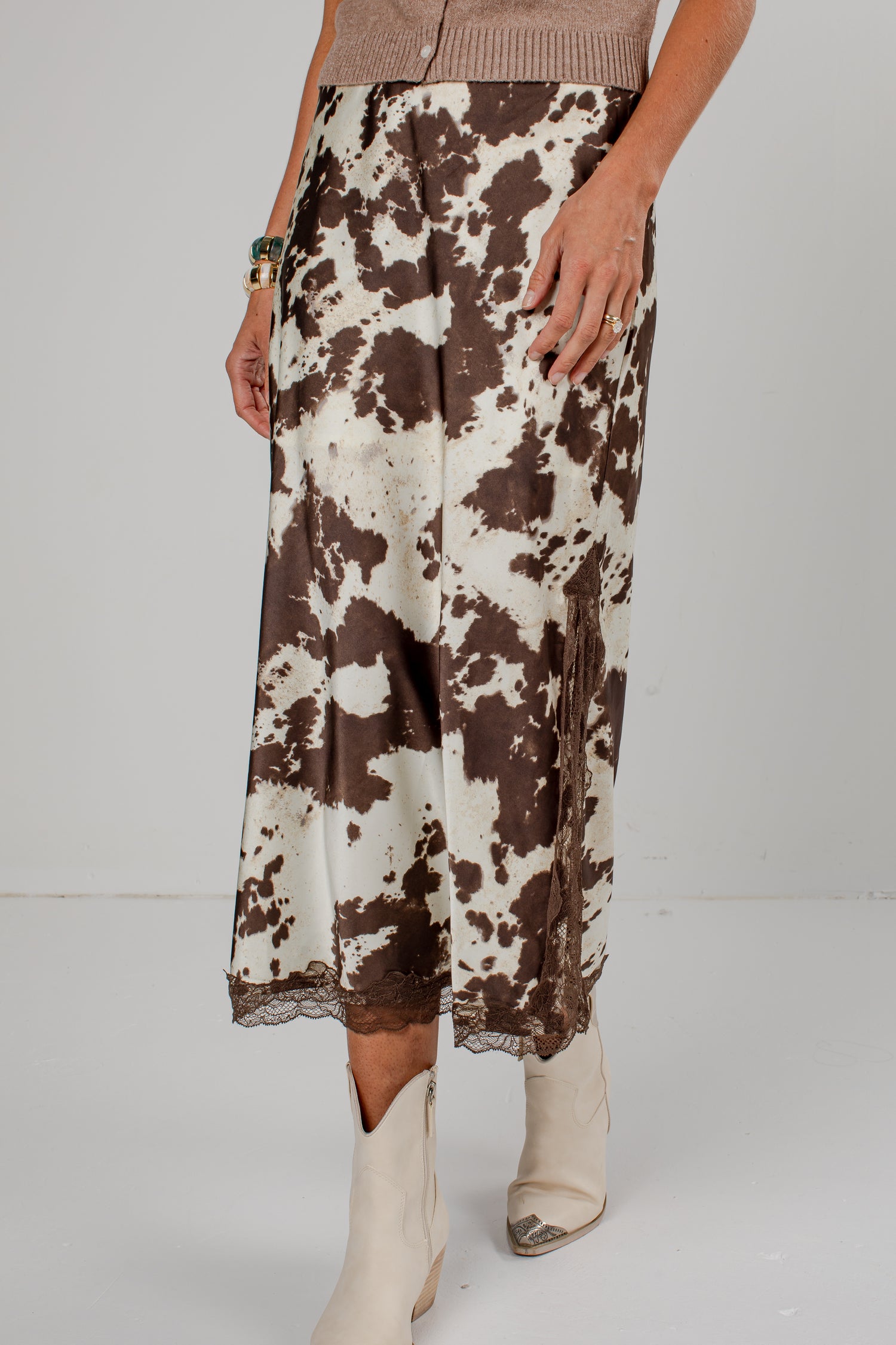 A model wearing a brown and cream cow-print midi skirt with a lace hem, paired with beige cowboy boots and a beige knit top, shown against a plain light gray studio background.