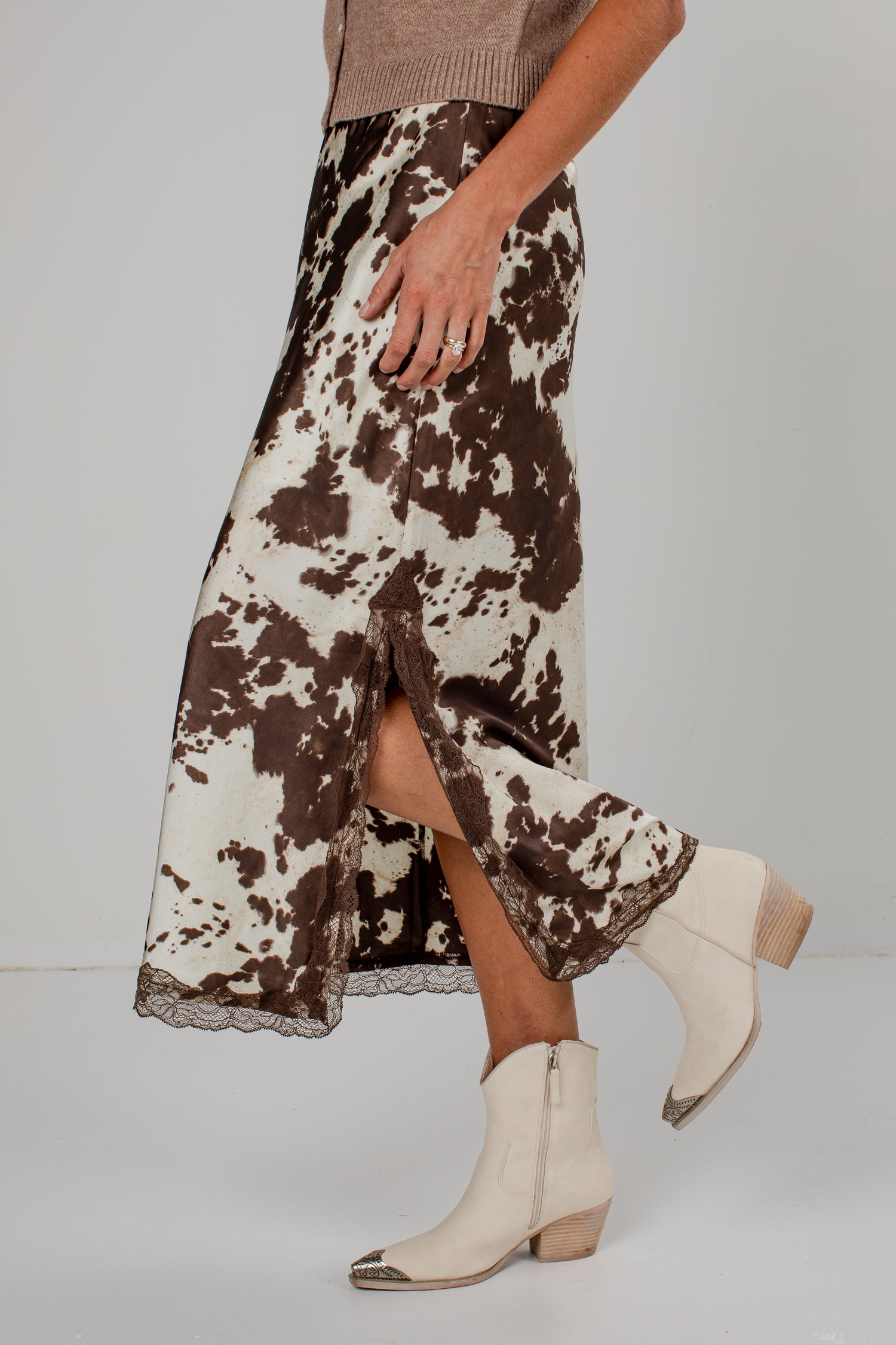 A model is shown from the waist down wearing a brown and white cow-print midi skirt with a lace hem, paired with cream-colored pointed-toe ankle boots.